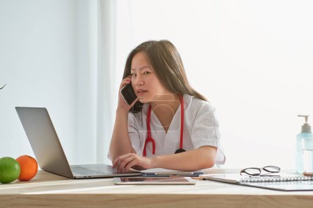 Photo for Focused Asian female physician in uniform talking on smartphone while working at table with laptop and documents in light clinic - Royalty Free Image
