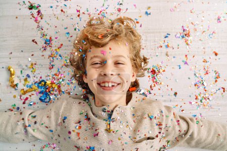 Téléchargez les photos : Joyeux garçon preteen aux cheveux bouclés blonds couché sur un sol stratifié léger recouvert de confettis multicolores et de paillettes - en image libre de droit