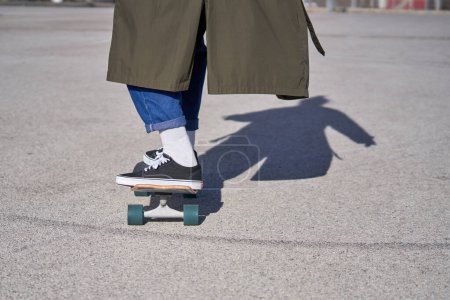 Photo for From below of side view of crop anonymous male in sneakers and long raincoat and jeans standing on skateboard on asphalt ground in park in sunny day - Royalty Free Image