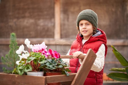 Photo for Cute smiling preschool boy in warm outerwear standing near wooden garden cart with blooming vivid flowers in pots growing in botanical garden - Royalty Free Image