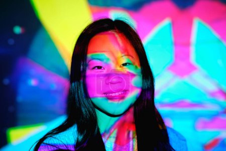 Foto de Alegre hembra asiática con pelo negro mirando a la cámara con sonrisa mientras está de pie cerca de la pared bajo brillantes luces de neón multicolores en el estudio - Imagen libre de derechos