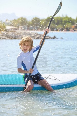 Photo for Full length of excited kid in swimsuit shouting while sitting on SUP board and rowing with paddle among rippling sea water during summer vacation - Royalty Free Image