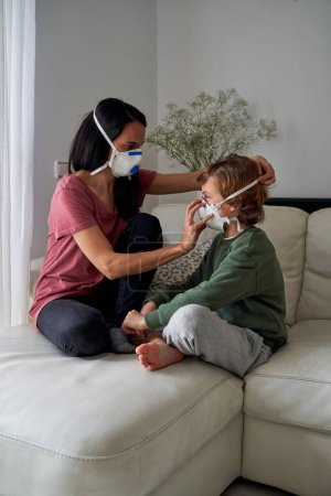 Photo for Side view of brunette and kid sitting on sofa together and putting on protective masks - Royalty Free Image
