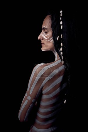 Téléchargez les photos : Jeune brune amérindienne avec des rayures blanches peintes sur le corps debout nu sur fond sombre tout en fermant les yeux - en image libre de droit
