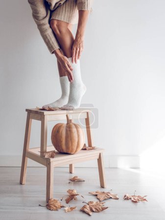 Photo for Crop unrecognizable woman wearing white socks standing on wooden stand decorated with pumpkin and dried autumn leaves against white background - Royalty Free Image