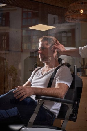 Téléchargez les photos : Homme confiant hipster assis dans un fauteuil et obtenir une nouvelle coupe de cheveux par coiffeur professionnel dans le salon de coiffure - en image libre de droit