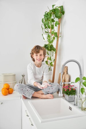 Photo for Full body of barefoot smiling boy wearing pajama holding bar of soap for cleaning routine near sink in morning in kitchen - Royalty Free Image
