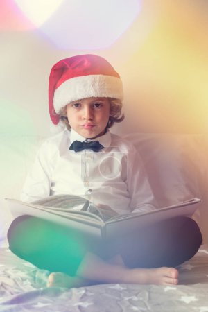 Foto de Cuerpo completo de chico descalzo en traje y sombrero de Santa mirando a la cámara mientras lee libro desinteresado en la cama durante la celebración de Navidad - Imagen libre de derechos