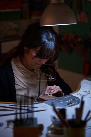 Photo for From above of focused adult ethnic female goldsmith sitting at workbench and repairing golden jewelry with saw in traditional workshop - Royalty Free Image