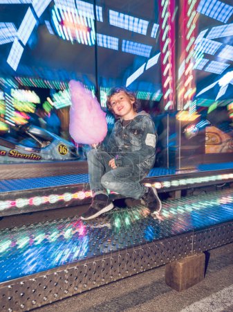 Foto de Cuerpo completo de niño alegre con dulces dulces de algodón mirando a la cámara mientras está sentado cerca de la atracción de automóviles de choque en el parque de atracciones - Imagen libre de derechos