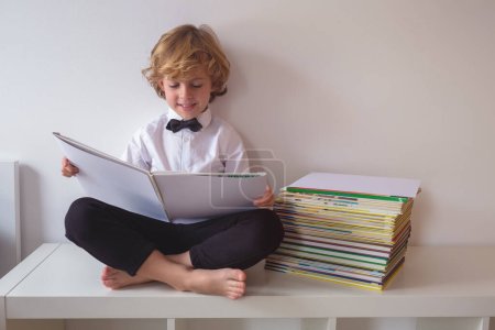 Photo for Full body of barefoot smiling boy in formal wear reading textbook while sitting on table with crossed legs on white background - Royalty Free Image