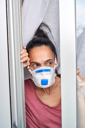 Photo for Woman with black hair in protective mask with valve looking away out opened window while standing in room during coronavirus pandemic - Royalty Free Image