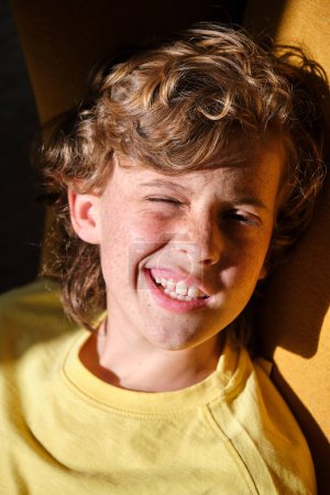 Foto de Alegre niño con pecas en ropa amarilla haciendo cara mientras mira a la cámara a la luz del sol - Imagen libre de derechos