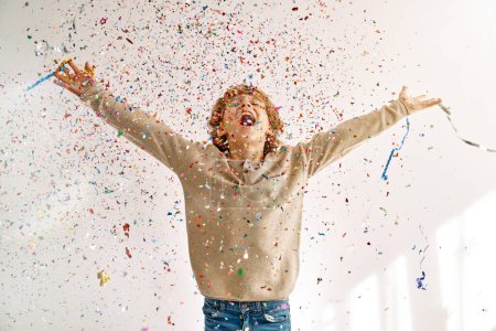 Photo for Excited preteen boy with curly hair rejoicing over surprise and screaming happily while standing under falling shiny spangles and confetti - Royalty Free Image