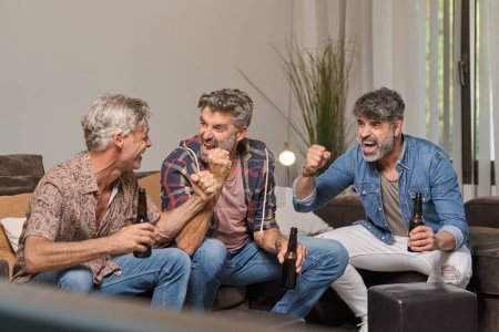 Téléchargez les photos : Les hommes adultes se regardent et célèbrent le but sur le match alors qu'ils sont assis sur le canapé et boivent de la bière à la maison - en image libre de droit