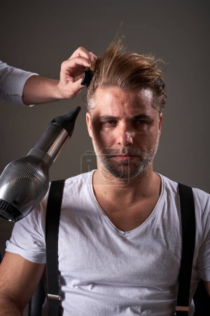 Téléchargez les photos : Homme adulte barbu confiant en vêtements décontractés et barbier méconnaissable séchant les cheveux du client et les peignant avec une brosse à cheveux - en image libre de droit
