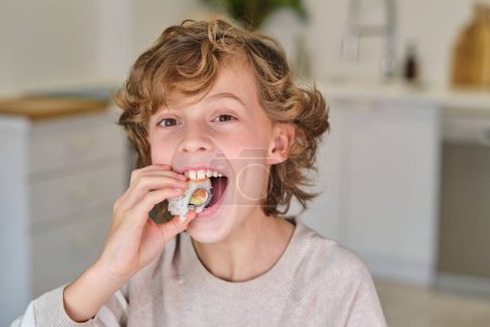 Foto de Adorable niño pequeño con el pelo rizado comiendo delicioso rollo y mirando a la cámara en la cocina en casa - Imagen libre de derechos