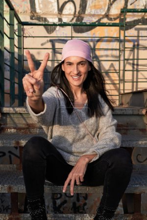 Téléchargez les photos : Femme hispanique souriante en foulard rose de la campagne de sensibilisation au cancer du sein assise sur un escalier en métal et montrant un geste de deux doigts en regardant la caméra - en image libre de droit