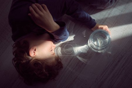 Photo for Overhead of little kid with curly hair lying peacefully on floor in sun ray shining through glass bottle with water - Royalty Free Image