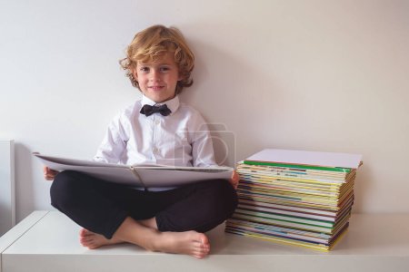 Photo for Full body of cheerful boy in formal wear looking at camera while reading textbook near pile of literature on white background - Royalty Free Image