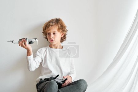 Photo for Isolated adorable curly child sitting against white background and looking at toy drone in hand - Royalty Free Image