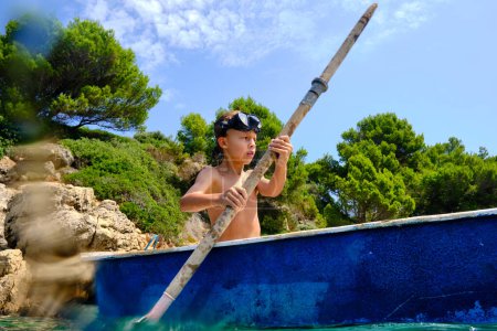 Téléchargez les photos : Garçon en masque de plongée bateau à rames avec pagaie sur la mer contre le rivage rocheux avec arbre pendant le voyage d'été en station tropicale - en image libre de droit