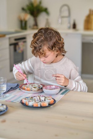 Téléchargez les photos : Garçon concentré avec les cheveux bouclés assis à la table en bois et regardant vers le bas tout en mangeant des rouleaux de sushi avec des baguettes dans la cuisine à la maison - en image libre de droit