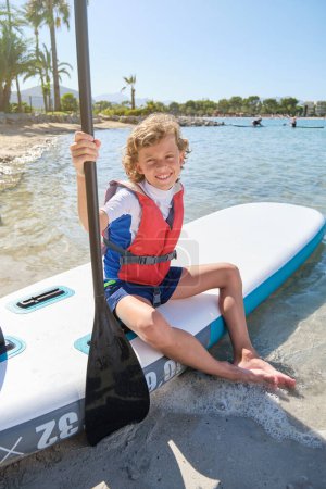 Téléchargez les photos : Pleine longueur de garçon pieds nus en maillot de bain et gilet de sauvetage orange souriant et regardant la caméra tout en étant assis sur la planche à pagaie sur la plage de sable pendant les vacances d'été - en image libre de droit