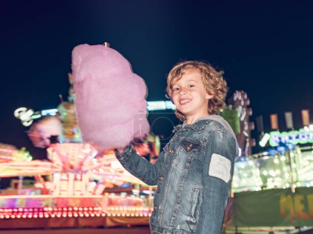 Téléchargez les photos : Garçon ravi avec bonbons en coton doux à la main regardant la caméra tout en se tenant près du carrousel lumineux dans le parc d'attractions en soirée - en image libre de droit