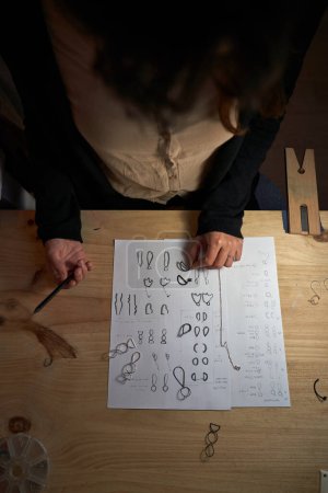 Foto de Desde arriba de la cosecha joyero anónimo con colgante se centra mientras que la creación de bocetos con lápiz en la mesa de madera en el taller oscuro - Imagen libre de derechos