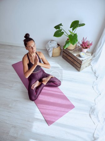 Photo for From above of female in activewear sitting on mat in Padmasana pose with namaste gesture and doing yoga with closed eyes in living room - Royalty Free Image