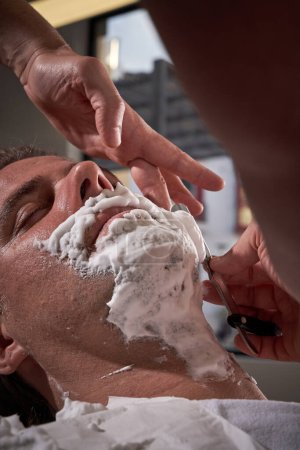 Photo for Crop anonymous skilled barber with razor shaving foamy beard of male client sitting with closed eyes during appointment in barbershop - Royalty Free Image