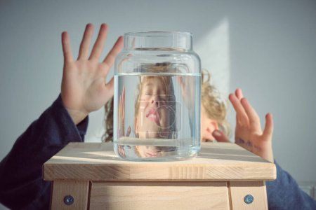 Photo for Frightened kid with blond hair and closed eyes raising arms and reflecting in glass transparent bottle of water placed on wooden stool against white background - Royalty Free Image