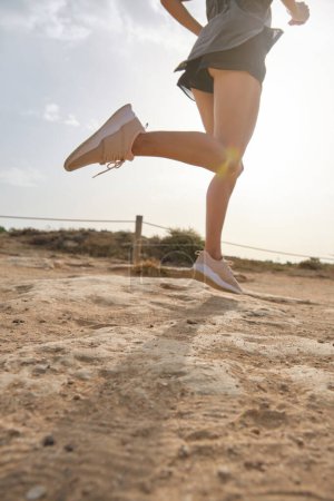 Foto de Crop atleta femenina en ropa deportiva y zapatillas de deporte que llevan un estilo de vida saludable y trotar en el camino de arena con arbustos en el día soleado - Imagen libre de derechos