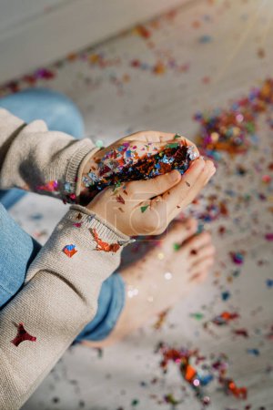 Foto de Alto ángulo de cultivo niño descalzo anónimo sentado en el suelo con confeti colorido en las manos - Imagen libre de derechos