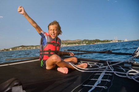 Foto de Cuerpo completo de niño alegre en chaleco salvavidas gritando y levantando el brazo con alegría mientras está sentado en un velero en ondulante mar azul - Imagen libre de derechos