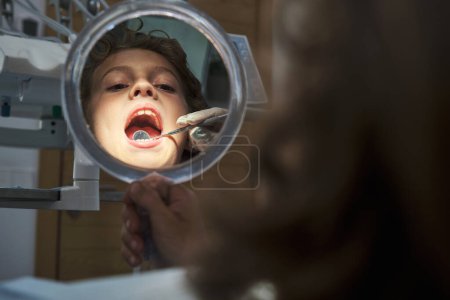 Foto de Cosecha dentista anónimo utilizando la herramienta para examinar los dientes del niño reflejándose en el espejo dental en la clínica moderna - Imagen libre de derechos