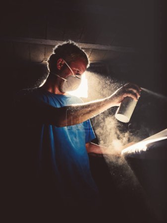 Photo for Male master in respirator spraying aerosol while dying surfboard in dark handicraft workshop lightened by sunlight shining through hole - Royalty Free Image