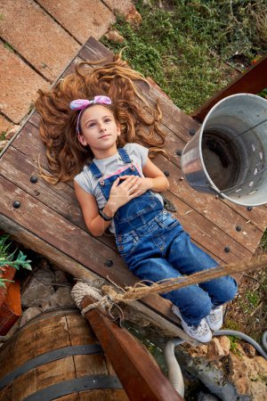 Photo for From above full body of cute girl in denim overall looking up with dreamy face while lying on wooden bench in countryside - Royalty Free Image
