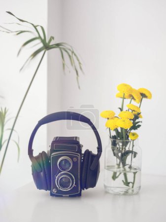 Photo for Musical headphones on vintage black photo camera placed on white table near vase with yellow flowers in room at home - Royalty Free Image
