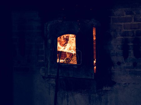 Photo for Black dirty furnace with bright burning flame built in brick wall covered with soot in dark glass factory - Royalty Free Image