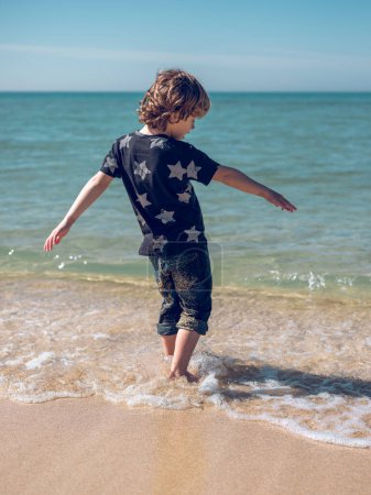 Foto de Vista trasera cuerpo completo de niño irreconocible caminando en aguas poco profundas en la playa de arena en el soleado día de verano - Imagen libre de derechos