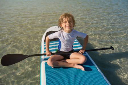 Photo for Full body portrait of smiling boy with paddle sitting on SUP board and looking at camera in middle of sea during summer vacation - Royalty Free Image