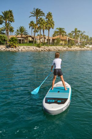 Photo for Back view of unrecognizable child standing on SUP board and paddling in sea with palm trees on beach on background during summer vacation - Royalty Free Image