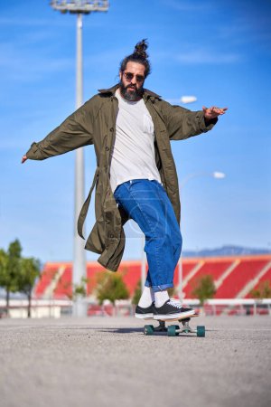 Photo for Low angle of full body delighted man in trendy coat and jeans wearing sunglasses riding stunt on longboard in park in sunny day - Royalty Free Image