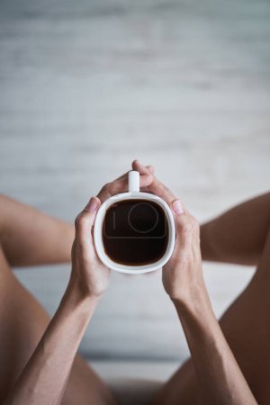 Photo for From above of anonymous female sitting with mug of americano in hands while having coffee break in room at home - Royalty Free Image
