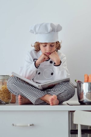 Photo for From below of full body cute preteen boy in chef suit and cap sitting on kitchen counter with crossed legs and reading cookbook attentively - Royalty Free Image