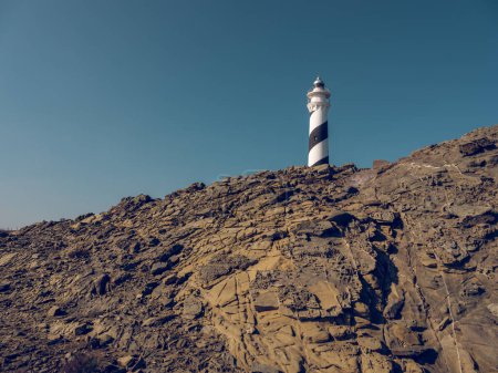 Photo for From below of white lighthouse located on top of rough rocky formations under cloudless blue sky - Royalty Free Image