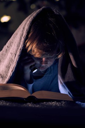 Foto de Enfocado chico leyendo interesante libro con linterna mientras está acostado en la cama debajo de cuadros calientes en la habitación oscura en la noche - Imagen libre de derechos