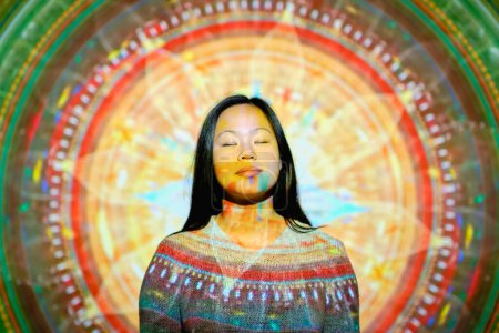 Téléchargez les photos : Tranquille asiatique femelle avec lumière sur le visage et les yeux fermés debout près du mur avec mandala lumineux avec ornement créatif et méditer en studio - en image libre de droit